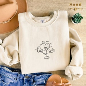 May include: A cream-colored sweatshirt with a black line drawing of Mickey Mouse juggling. The sweatshirt is laid out on a wooden surface with a brown hat, blue jeans, and a small potted plant.