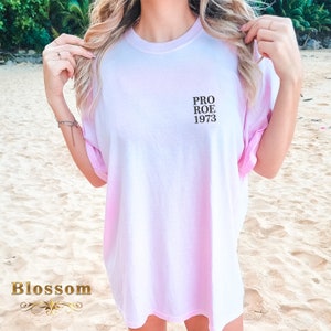 May include: A light pink t-shirt with the text "PRO ROE 1973" printed in black. The shirt has a gradient effect, fading from white to pink. The word "Blossom" is printed in gold at the bottom left.