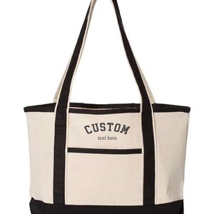May include: A large, beige canvas tote bag with black handles and trim. The bag has a front pocket and the text "CUSTOM text here" is embroidered on the front.
