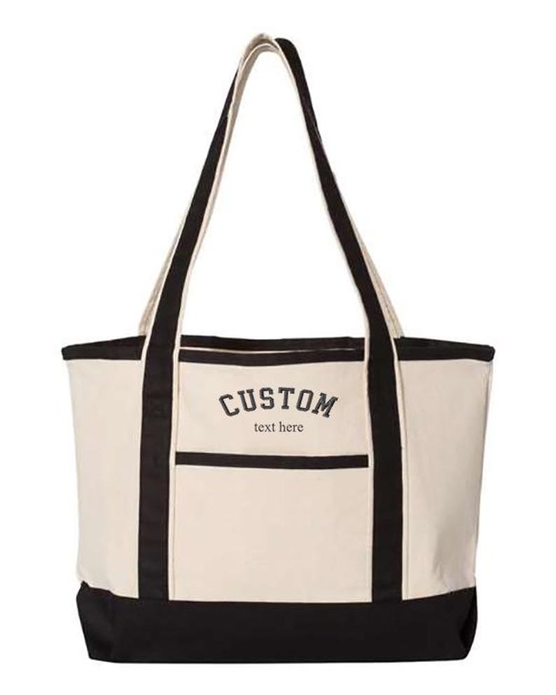 May include: A large, beige canvas tote bag with black handles and trim. The bag has a front pocket and the text "CUSTOM text here" is embroidered on the front.
