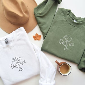 May include: Two sweatshirts with a simple line drawing of Mickey Mouse on them. One sweatshirt is white and the other is green.