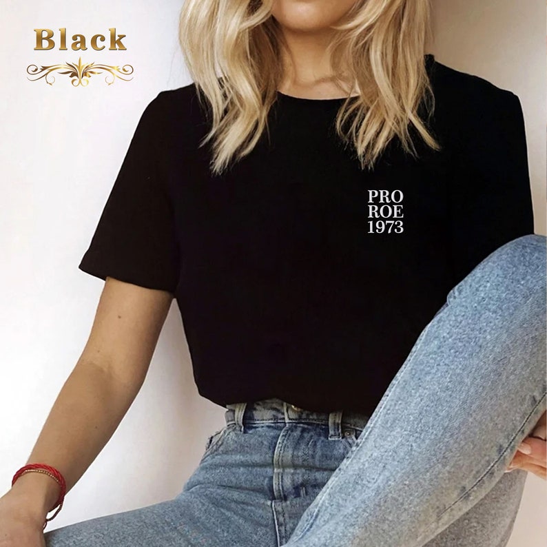 May include: Black t-shirt with the text "PRO ROE 1973" in white. The shirt has short sleeves and a crew neck. The word "Black" is displayed in gold script in the upper left corner.