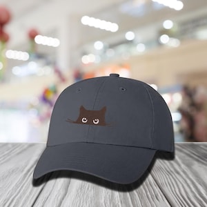 May include: A dark gray baseball cap featuring an embroidered brown cat face peeking over the brim. The cat's eyes are white. The cap is made of a soft material and has a curved brim.