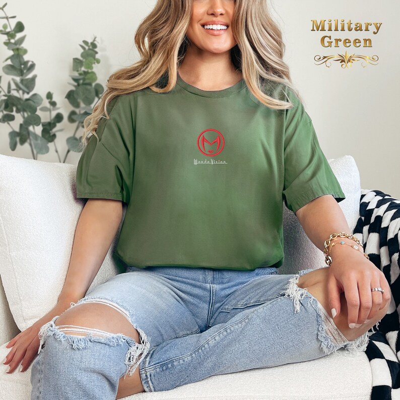 May include: A woman wearing a military green t-shirt with a red "M" logo and the text "Made Visine" on the front. She is sitting on a white sofa with her legs crossed.