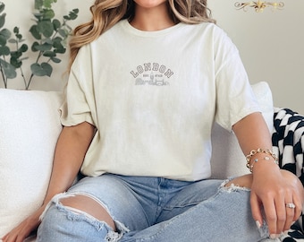 May include: A woman wearing a light beige t-shirt with the text "LONDON EST. 47AD" and a graphic of the London skyline. She is wearing blue jeans with rips in the knees.