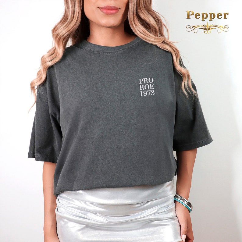 May include: Dark gray t-shirt with the text "PRO ROE 1973" in white. The shirt has short sleeves and a relaxed fit. The shirt is made of a soft material.