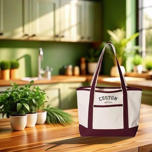 May include: A white canvas tote bag with maroon trim and handles. The bag has a pocket on the front with the text "CUSTOM text here" embroidered in gray.