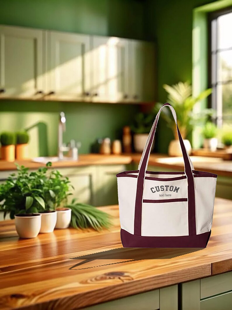 May include: A white canvas tote bag with maroon trim and handles. The bag has a pocket on the front with the text "CUSTOM text here" embroidered in gray.