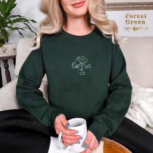 May include: A dark green sweatshirt with a white outline of Mickey Mouse on the front. The sweatshirt is being worn by a person sitting on a couch.