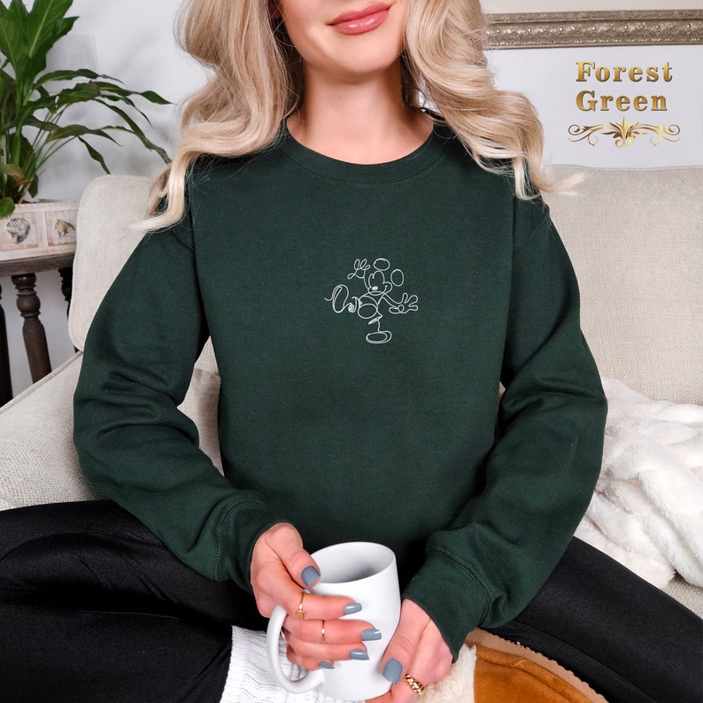 May include: A dark green sweatshirt with a white outline of Mickey Mouse on the front. The sweatshirt is being worn by a person sitting on a couch.