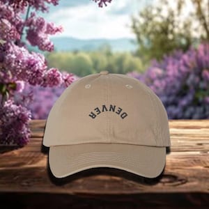 May include: A tan baseball cap with the word "DENVER" arched across the front in dark gray lettering. The cap has a curved brim and a classic six-panel design. The background is a blurred outdoor scene.