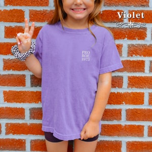 May include: A violet-colored t-shirt with the text "PRO ROE 1973" printed on the front. The shirt is short-sleeved and appears to be made of a soft material. The word "Violet" is written in a decorative font.