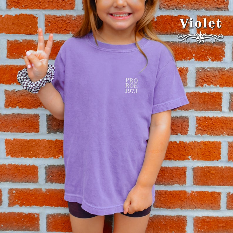 May include: A violet-colored t-shirt with the text "PRO ROE 1973" printed on the front. The shirt is short-sleeved and appears to be made of a soft material. The word "Violet" is written in a decorative font.