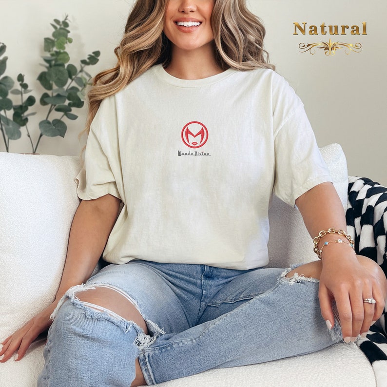 May include: A woman wearing a white t-shirt with a red WandaVision logo and the text "WandaVision". She is wearing blue jeans and sitting on a white sofa.