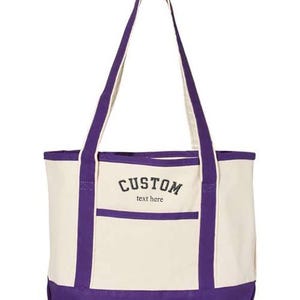 May include: A purple and white tote bag with a front pocket. The bag has a black embroidered design that says "CUSTOM text here".