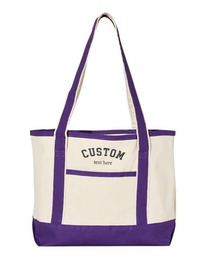 May include: A purple and white tote bag with a front pocket. The bag has a black embroidered design that says "CUSTOM text here".