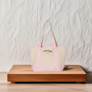 May include: A pink and beige canvas tote bag with a front pocket and the text "CUSTOM text here" printed on the front.