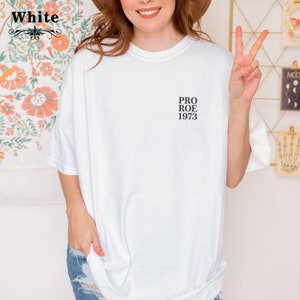 May include: White t-shirt with the text "PRO ROE 1973" printed on the front. The shirt has a relaxed fit and short sleeves. The word "White" is printed on the top left corner of the image.