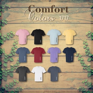 Op de afbeelding: Een display van Comfort Colors t-shirts in diverse kleuren, waaronder roze, zwart, blauw en geel. Elk shirt is gelabeld met zijn kleurnaam, zoals "blossom" en "black". De shirts zijn gerangschikt op een houten achtergrond.