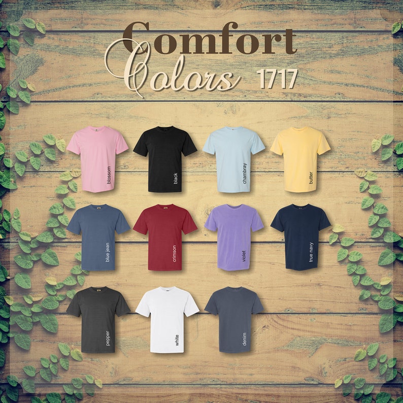 Op de afbeelding: Een display van Comfort Colors t-shirts in diverse kleuren, waaronder roze, zwart, blauw en geel. Elk shirt is gelabeld met zijn kleurnaam, zoals "blossom" en "black". De shirts zijn gerangschikt op een houten achtergrond.