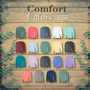 Op de afbeelding: Een display van Comfort Colors 1566 sweatshirts in diverse kleuren, waaronder blauwe spar, watermeloen, blauwe jeans, wit en krijtmunt. De afbeelding staat tegen een houten achtergrond met groene wijnranken.