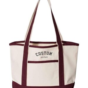 May include: A burgundy and cream canvas tote bag with a large pocket on the front. The bag has a custom embroidered design that says "CUSTOM text here".
