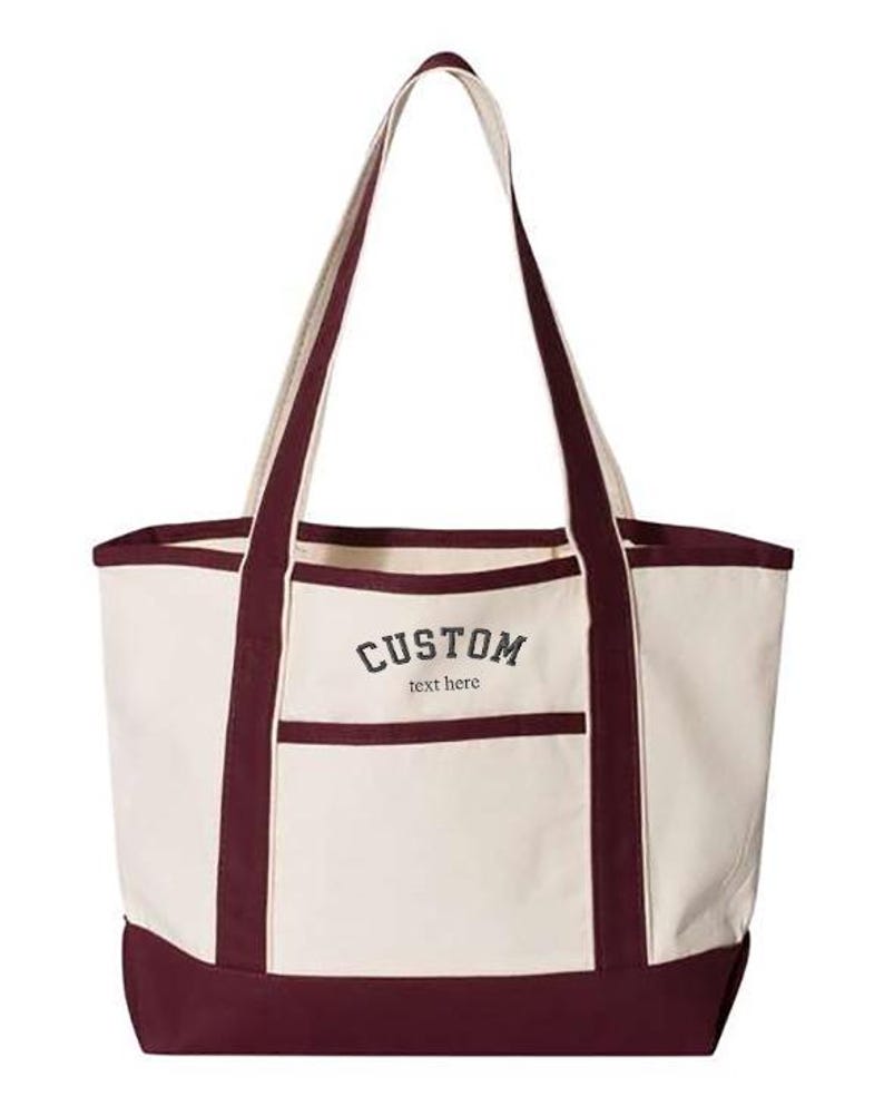 May include: A burgundy and cream canvas tote bag with a large pocket on the front. The bag has a custom embroidered design that says "CUSTOM text here".
