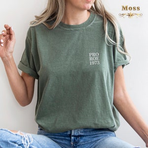 May include: Olive green t-shirt with rolled-up sleeves. The shirt features the text "PRO ROE 1973" in white, centered on the chest. The shirt has a slightly faded appearance.