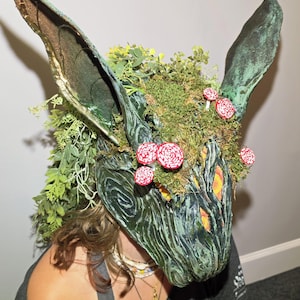 Könnte beinhalten: Eine dunkelgrüne, texturierte Tiermaske mit hohen, spitzen Ohren, verziert mit grünem Blattwerk, Moos und kleinen rot-weiß gepunkteten Pilzen. Die Maske weist tiefe, wirbelnde Muster und orangefarbene Augenöffnungen auf.