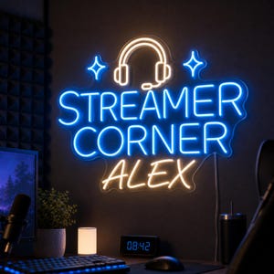 May include: A custom neon sign featuring a glowing white headphone icon flanked by two four-pointed stars above the blue neon text "STREAMER CORNER" and the name "ALEX" in warm yellow neon, mounted on a dark wall above a desk setup.