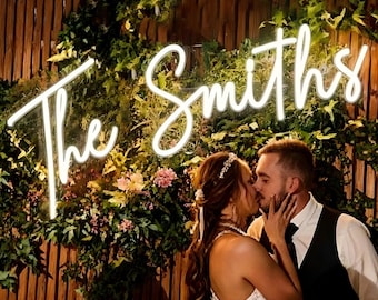 May include: A neon sign that reads "The Smiths" in a cursive font, mounted on a wall of greenery and wood. A couple is kissing in front of the sign, celebrating a wedding.