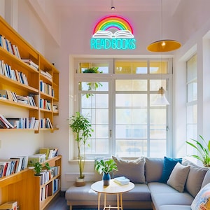 May include: A cozy reading nook with a neon sign that reads "READ BOOKS" above an open book and rainbow. Bookshelves line the wall, and a gray sectional sofa sits near a large window. A small table holds a vase and books.