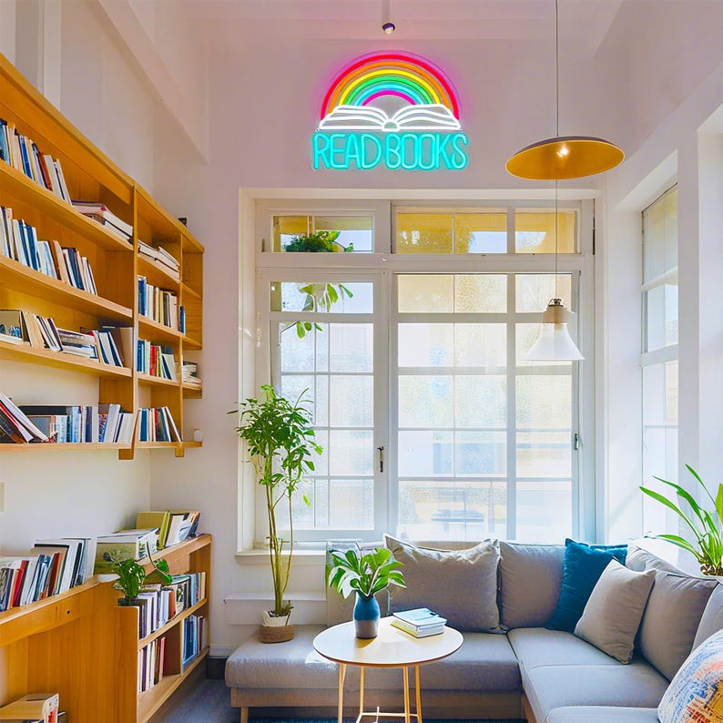 May include: A cozy reading nook with a neon sign that reads "READ BOOKS" above an open book and rainbow. Bookshelves line the wall, and a gray sectional sofa sits near a large window. A small table holds a vase and books.