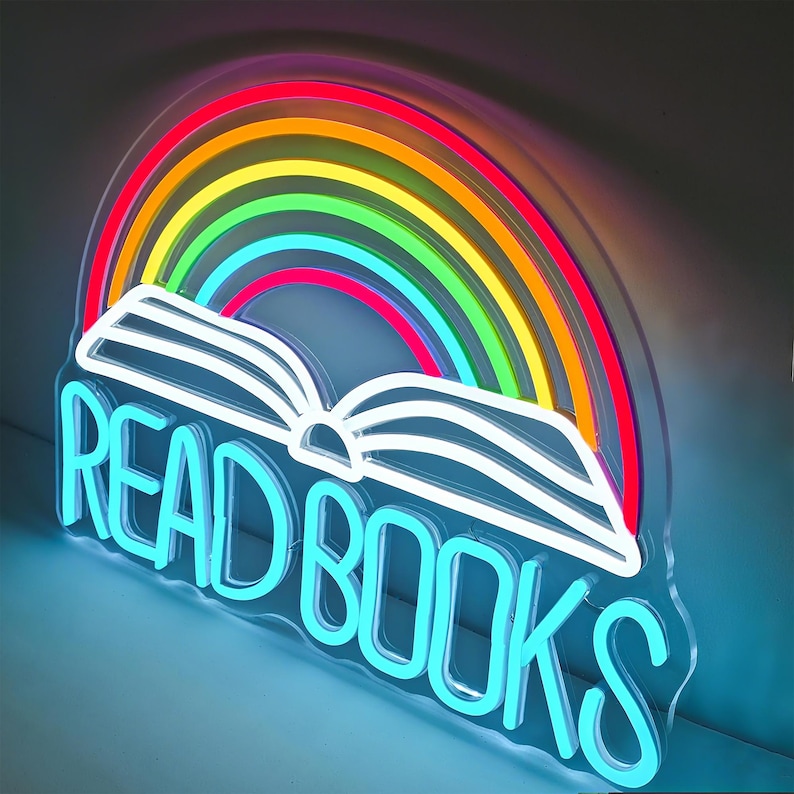 May include: A neon sign featuring a rainbow arching over an open book. The sign is outlined in white and has the words "READ BOOKS" in bright blue neon. The rainbow displays the colors red, orange, yellow, green, and blue.