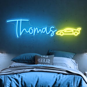 May include: A neon sign in blue and yellow illuminates a bedroom wall. The sign spells "Thomas" in cursive, followed by a car silhouette. The room features a bed with blue bedding and pillows, creating a modern aesthetic.