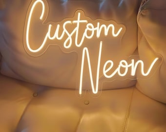 May include: A custom neon sign featuring the words Custom Neon in a glowing warm white script font. The sign is mounted on a clear acrylic backing that follows the contour of the lettering, creating a bright and illuminated display.
