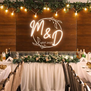 Può includere: Allestimento per ricevimento di nozze con sfondo in legno con insegna al neon con le lettere "M&D" e la data "EST.2025". Il tavolo è decorato con fiori, verde e luci a corda, creando un'atmosfera elegante.