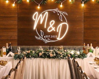 May include: A wedding reception setting with a wooden backdrop featuring a glowing neon sign with the letters "M&D" and the date "EST.2025". The table is decorated with flowers, greenery, and string lights, creating an elegant atmosphere.