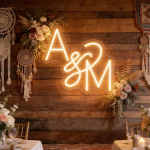 Pode incluir: Uma placa de néon que exibe as letras "A & M" em uma fonte cursiva, iluminada com um brilho quente e convidativo. A placa está colocada contra um fundo rústico de madeira, adornado com arranjos florais, criando um ambiente romântico.