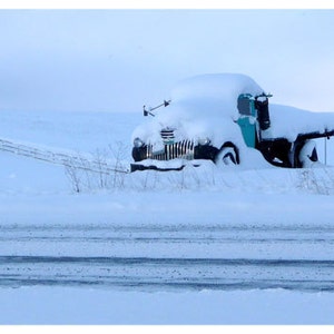 May include: A teal vintage truck is partially buried in a snowdrift. The truck is covered in snow, with only the front end and a portion of the bed visible. The truck is parked in a snowy field with a wooden fence in the background.