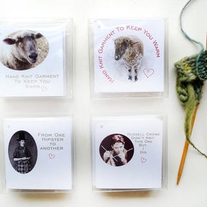 May include: Four white cards with black text and pink hearts. The cards are in a grid pattern. The top left card says "Hand Knit Garment To Keep You Warm". The top right card says "Hand Knit Garment To Keep You Warm". The bottom left card says "From One Hipster To Another". The bottom right card says "Russell Crowe Didn't Knit This One But I Did".