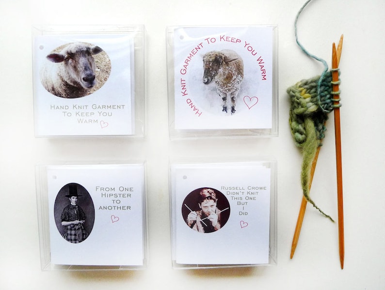 May include: Four white cards with black text and pink hearts. The cards are in a grid pattern. The top left card says "Hand Knit Garment To Keep You Warm". The top right card says "Hand Knit Garment To Keep You Warm". The bottom left card says "From One Hipster To Another". The bottom right card says "Russell Crowe Didn't Knit This One But I Did".