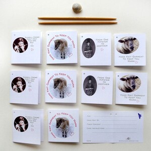 May include: Three white cards with a sheep illustration and text that reads "Hand Knit Garment to Keep You Warm."  The text is in pink and the cards have a hole punched in the top left corner.  The cards are on a white surface with two wooden knitting needles and a small white shell.