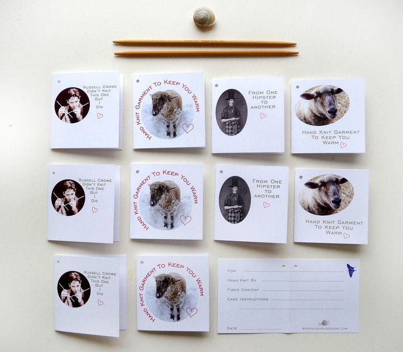 May include: Three white cards with a sheep illustration and text that reads "Hand Knit Garment to Keep You Warm."  The text is in pink and the cards have a hole punched in the top left corner.  The cards are on a white surface with two wooden knitting needles and a small white shell.