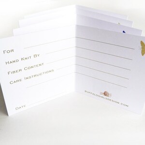 May include: A white card with gold lettering and illustrations. The card has lines for writing information about a knitted item, including "For", "Hand Knit By", "Fiber Content", "Care Instructions", and "Date". The card also features a small illustration of a butterfly and a knitting needle.