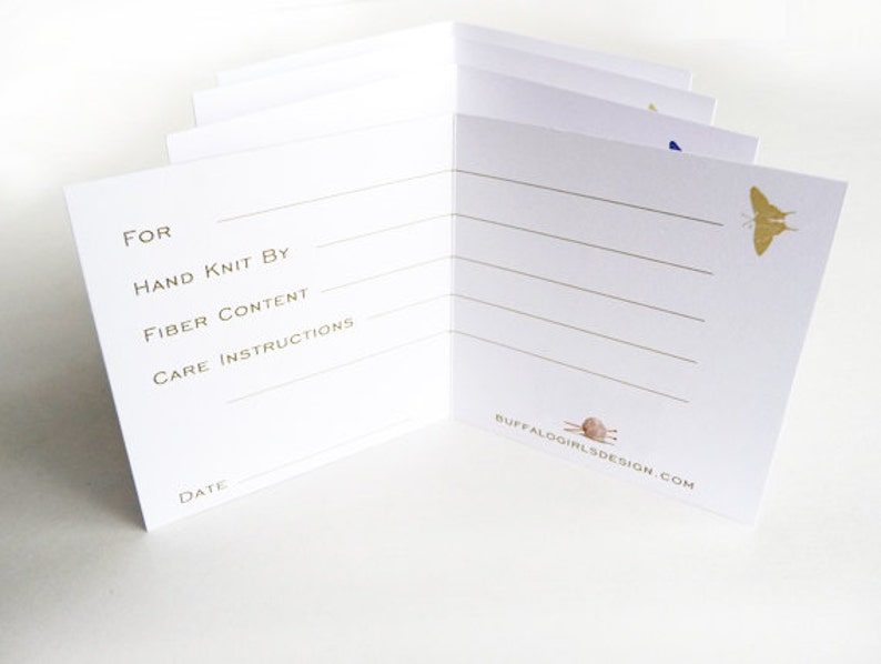 May include: A white card with gold lettering and illustrations. The card has lines for writing information about a knitted item, including "For", "Hand Knit By", "Fiber Content", "Care Instructions", and "Date". The card also features a small illustration of a butterfly and a knitting needle.