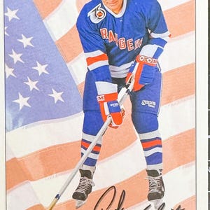May include: A hockey trading card featuring a man in a blue, red, and white New York Rangers jersey, holding a hockey stick. The background shows an American flag. The card is signed "Andy Bathgate".