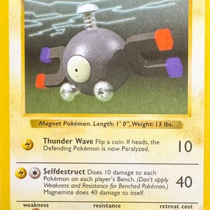 May include: A yellow Pokémon trading card featuring Magnemite, a gray, spherical creature with magnets. The card includes text about Thunder Wave and Selfdestruct attacks, with a weight of 13 lbs. and a length of 1' 0".