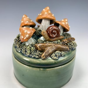 May include: A green ceramic trinket box with a lid featuring a snail crawling past a cluster of orange and white mushrooms with brown branches.