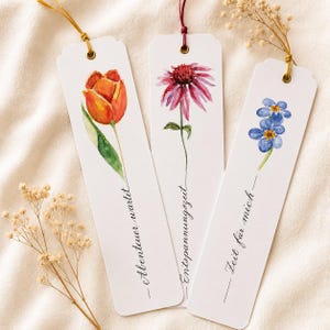 Könnte beinhalten: Drei rechteckige Lesezeichen aus weißem Papier mit abgerundeten oberen Ecken und Metallösen. Jedes zeigt eine Aquarell-Blumenillustration: eine orangefarbene Tulpe, einen rosa Sonnenhut und blaue Vergissmeinnicht mit deutschem Text.